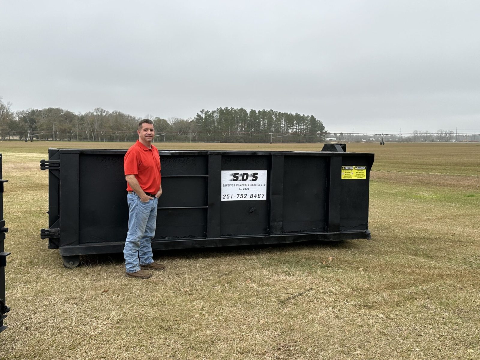A man in a red shirt and jeans stands next to a large black dumpster on a grassy field under an overcast sky.