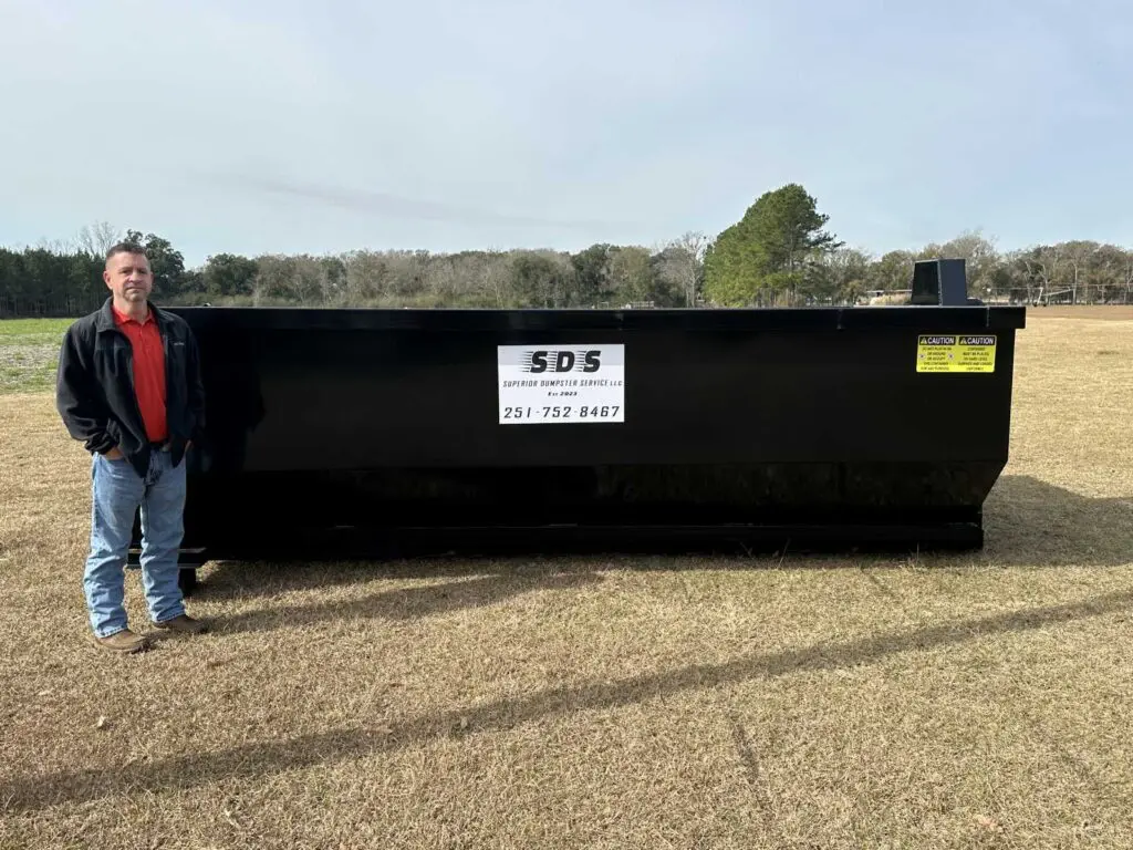 A man stands next to a large black dumpster with SDS Supreme Dumpsters Service signage on a grassy field.