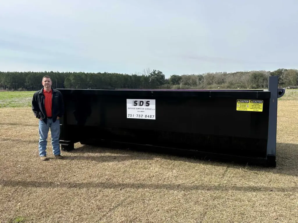 A man stands next to a large black dumpster on a grassy field with trees in the background.