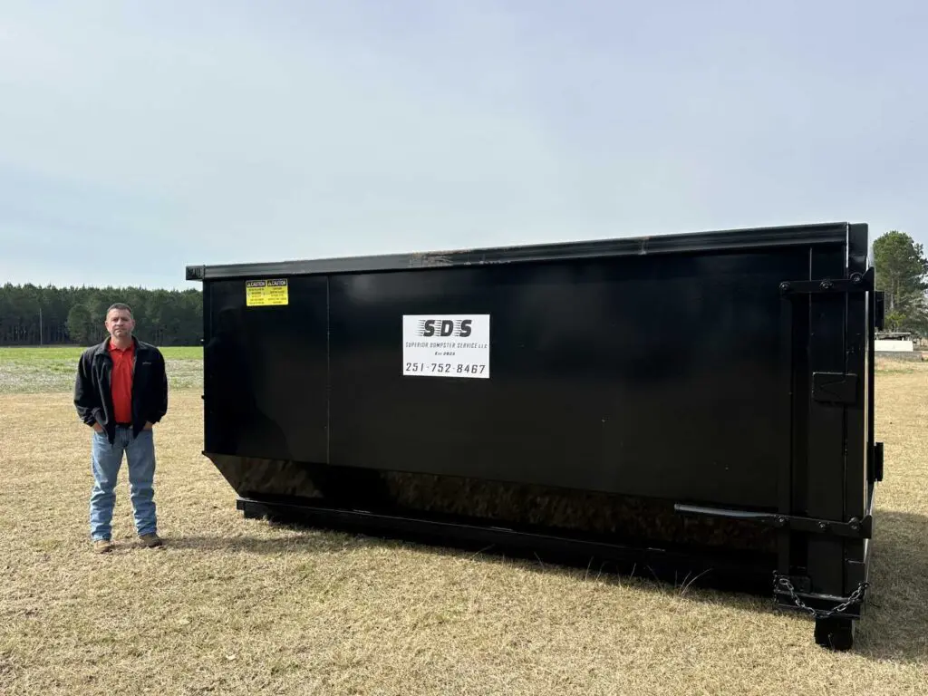 A man stands outdoors next to a large black dumpster labeled SDS on a grassy field with trees in the background.