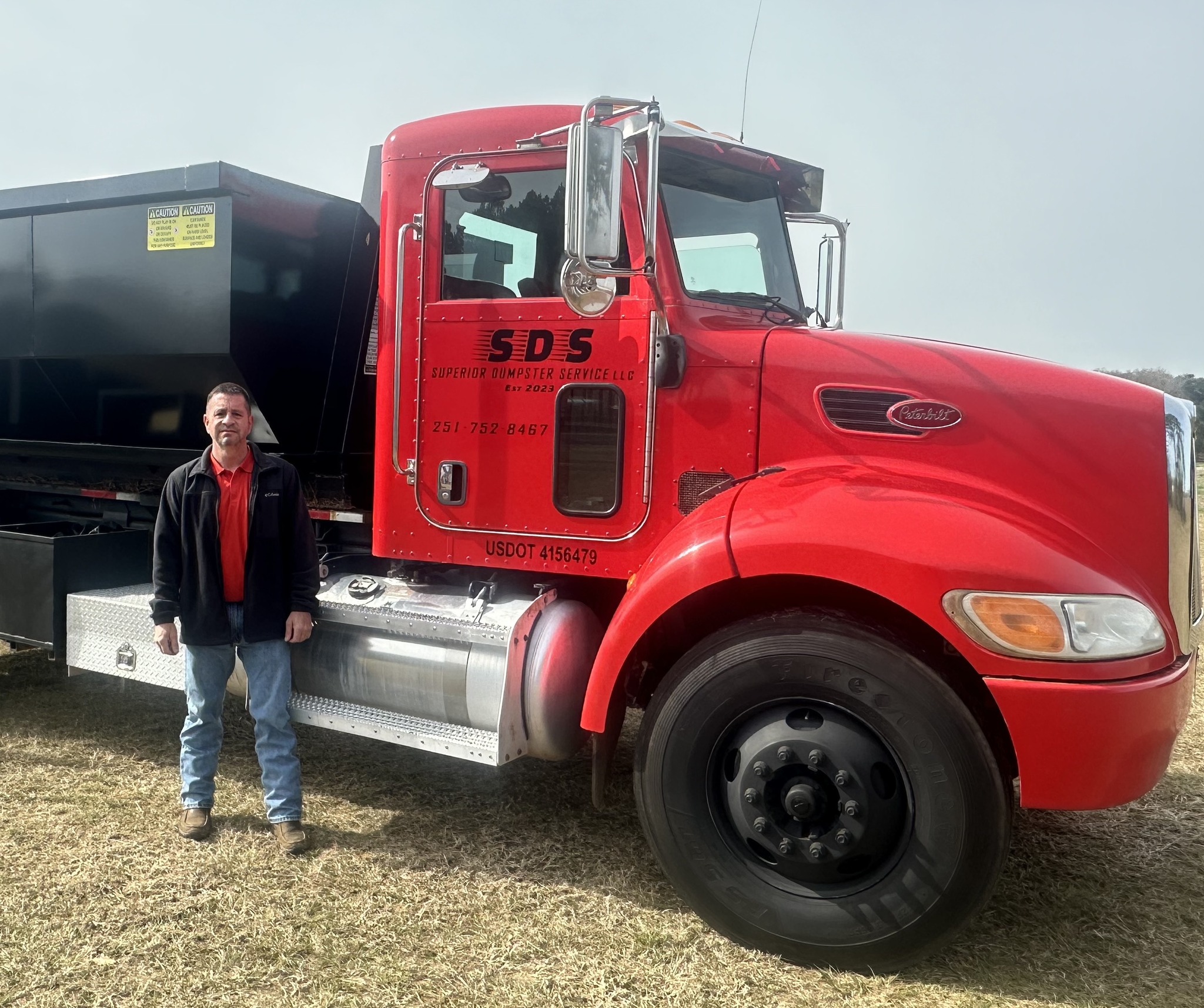 A man stands next to a large red truck with SDS and company details printed on the driver's door, parked on grass.