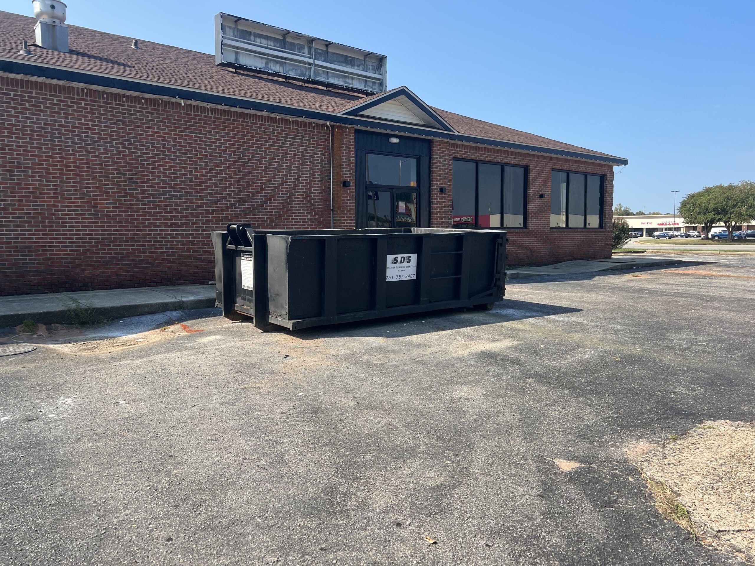 A large black commercial dumpster sits in the parking lot outside a red brick building with dark windows under a clear blue sky, highlighting the convenience of commercial dumpster rentals.