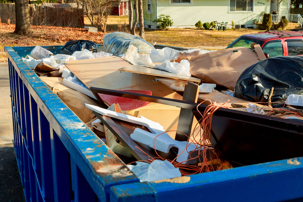 A large blue dumpster filled with various types of debris and garbage bags sits outdoors in a residential area.