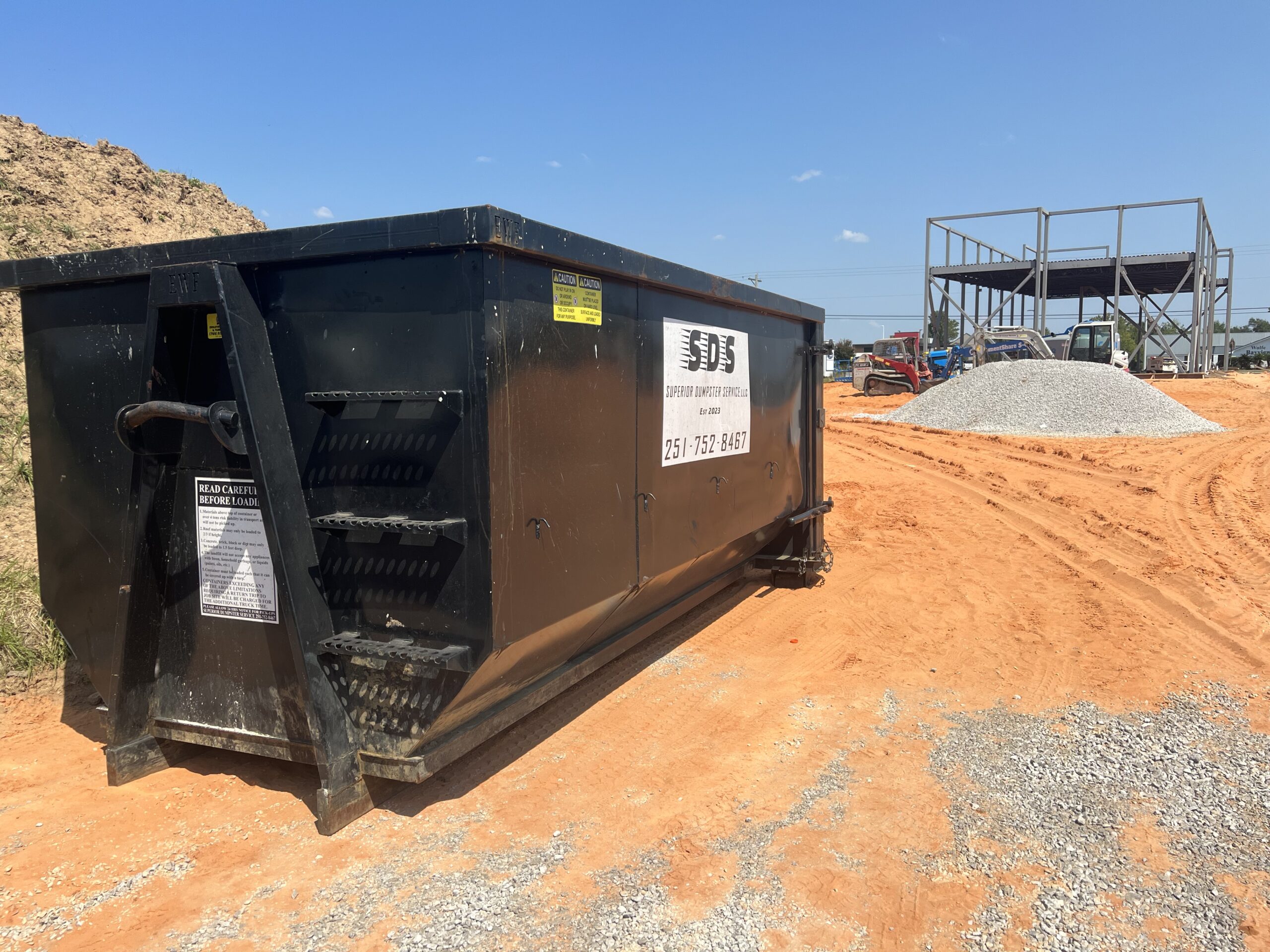 A large black contractor dumpster rentals unit sits on an orange dirt construction site near a gravel pile and partially constructed metal framework.