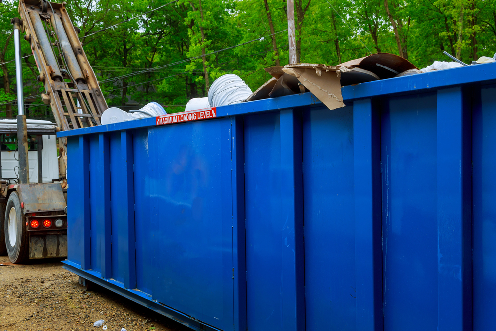 A large blue dumpster filled with construction debris and trash is positioned outdoors near a truck, with trees visible in the background.