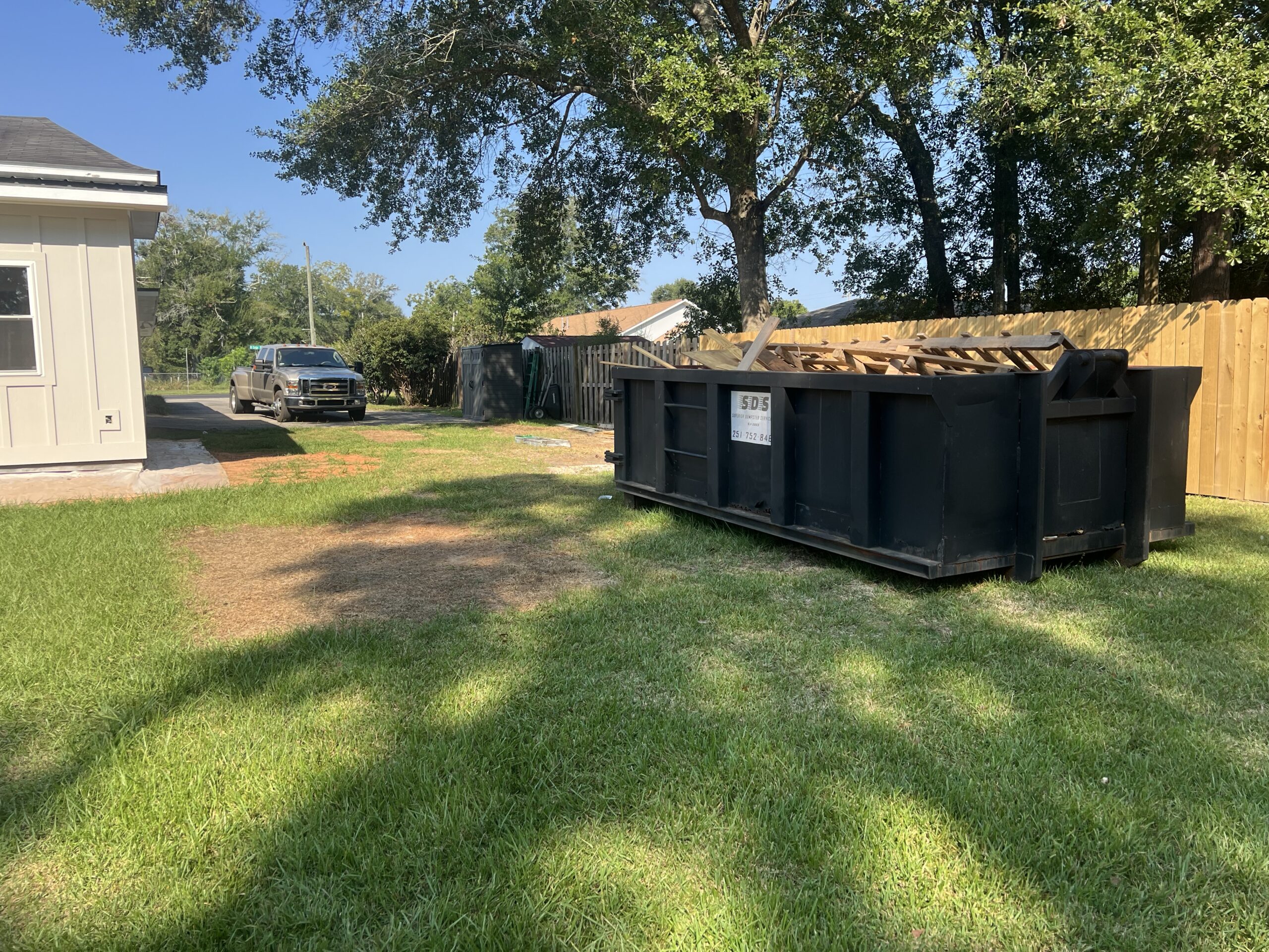 A black new construction dumpster sits filled with debris on grass near a wooden fence in a residential backyard; a house and parked truck are visible in the background.