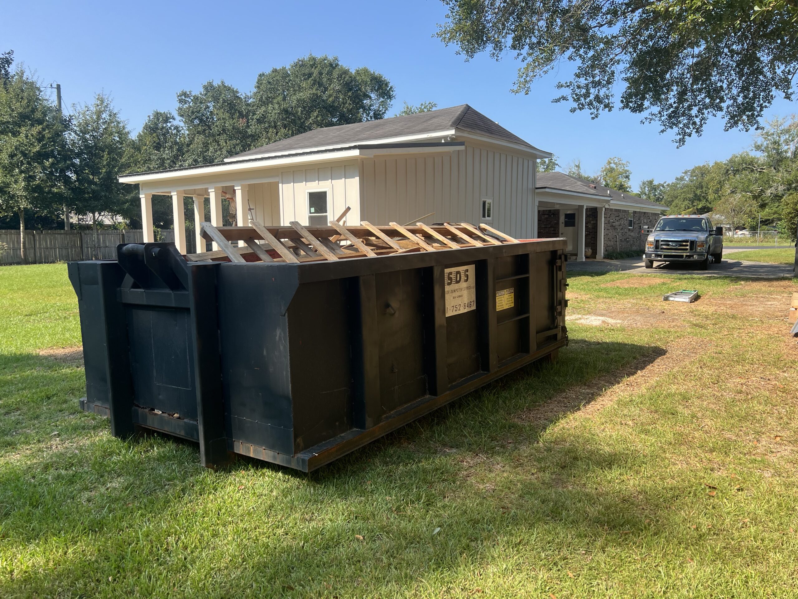 A large black new construction dumpster rental with wooden debris sits on grass in front of a beige house on a sunny day. A black truck is parked near the driveway in the background.