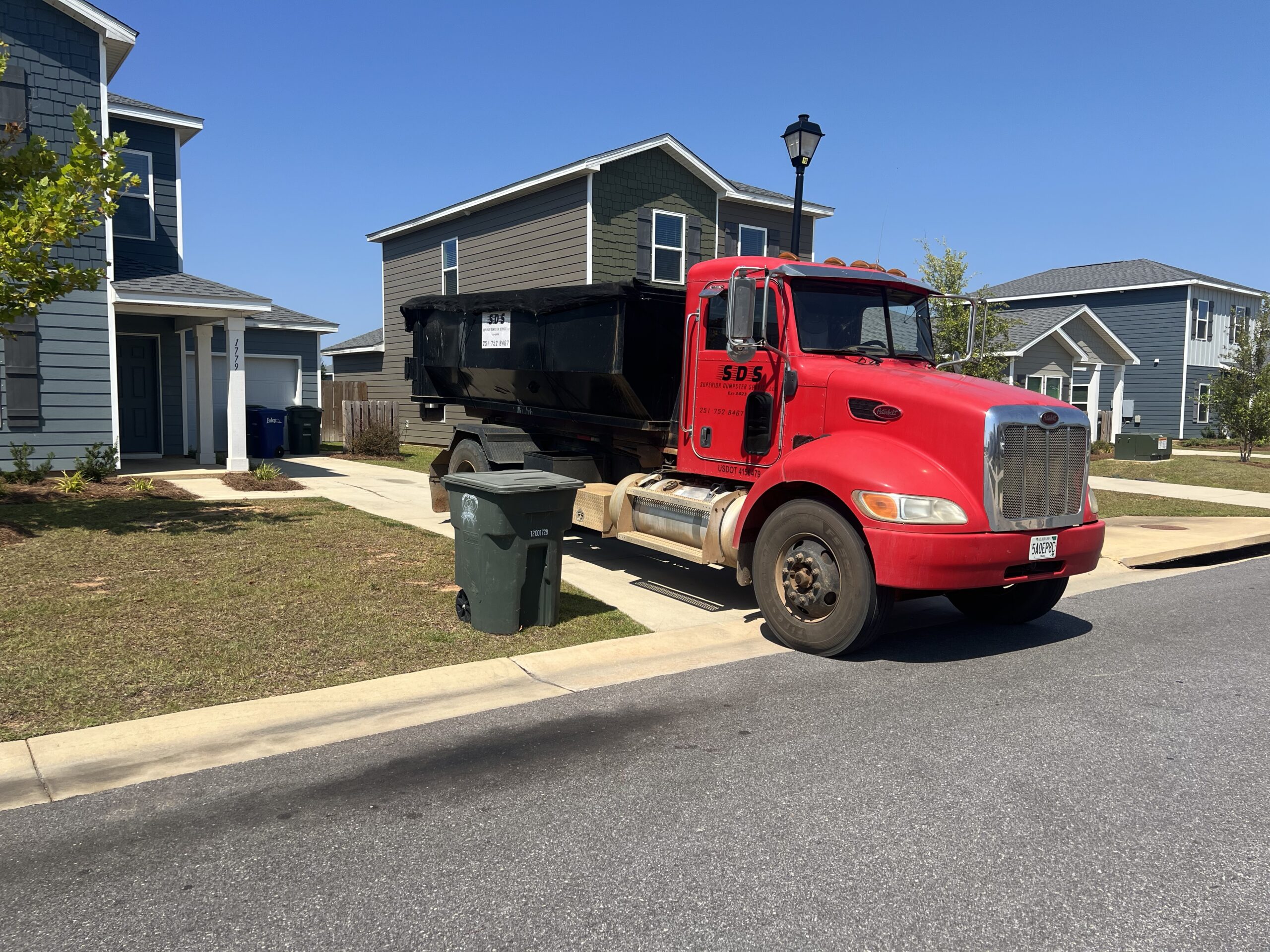 A red dump truck is parked on the street in a residential neighborhood near green and black garbage bins, suggesting residential dumpster rentals are available in the area.