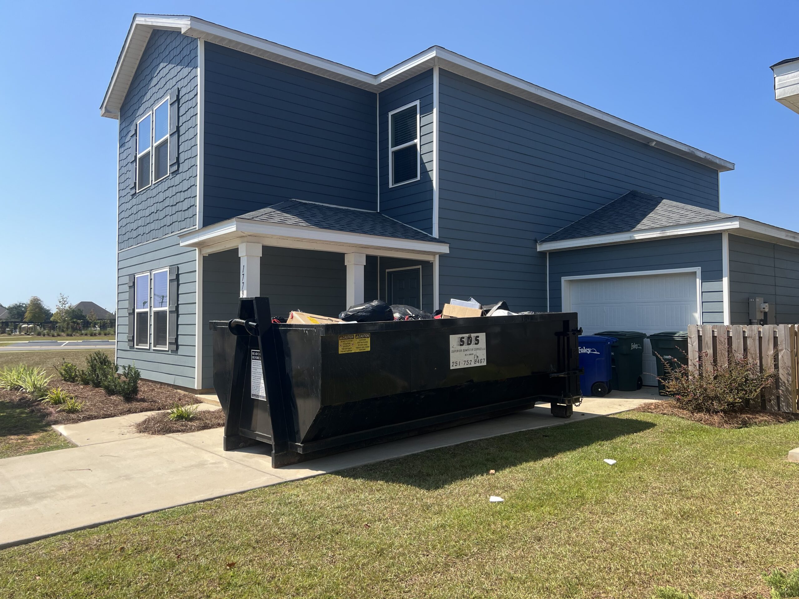 A large black yard waste dumpster rental sits in the driveway of a blue two-story house with various trash and debris inside.