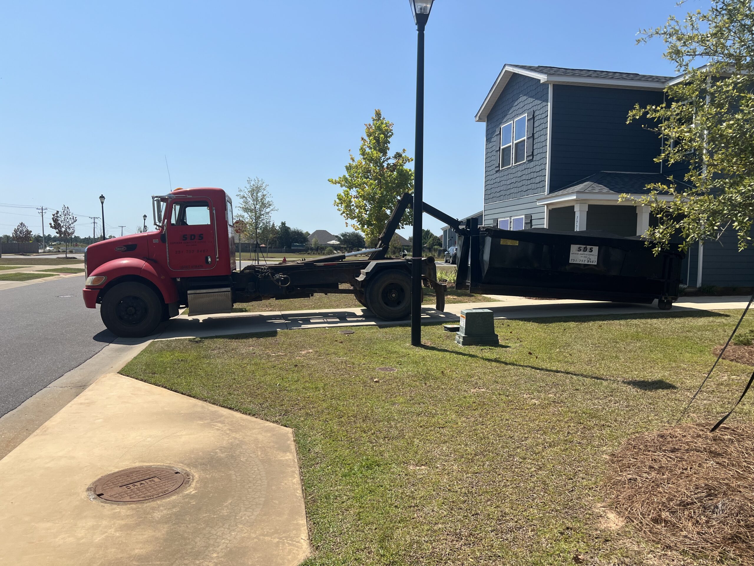 A red truck is delivering a large black yard waste dumpster rental onto the driveway of a two-story blue house on a sunny day.